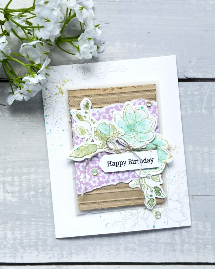 Handmade “Happy Birthday” card featuring a layered design with soft mint and green floral images, a lilac patterned paper panel, and textured kraft paper strips. The flowers are lightly watercoloured and tied with natural twine, with subtle ink splatter and small iridescent embellishments, photographed on a light wooden surface with white flowers above.