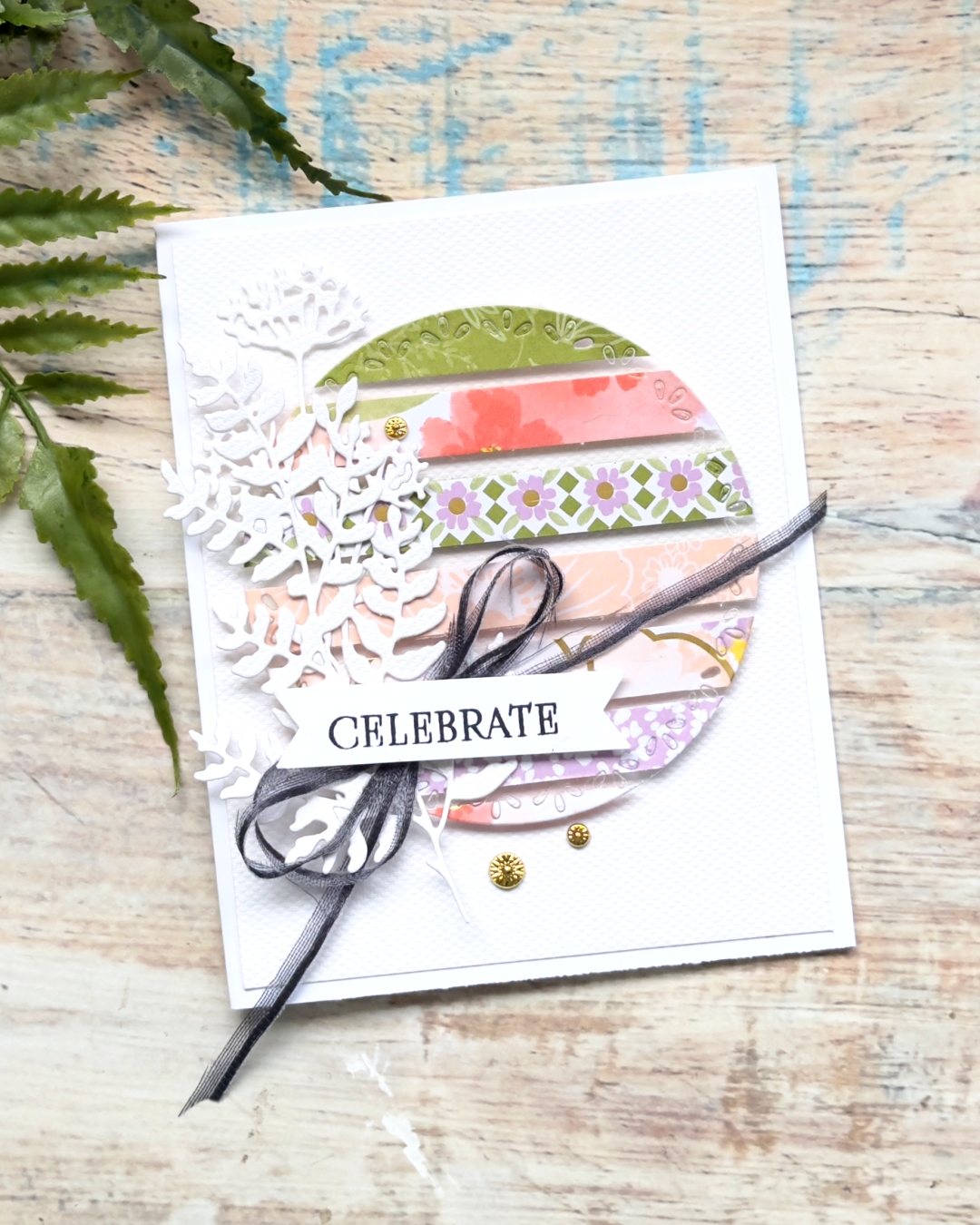 Handmade card using the floating technique with designer paper strips, circle die cut, white foliage die cuts, black ribbon and Celebrate sentiment.