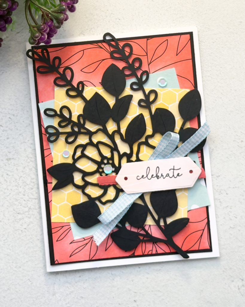Bold handmade “Celebrate” card created with blended coral background, layered patterned papers, black floral die cuts, soft blue ribbon and layered sentiment tag with iridescent embellishments.