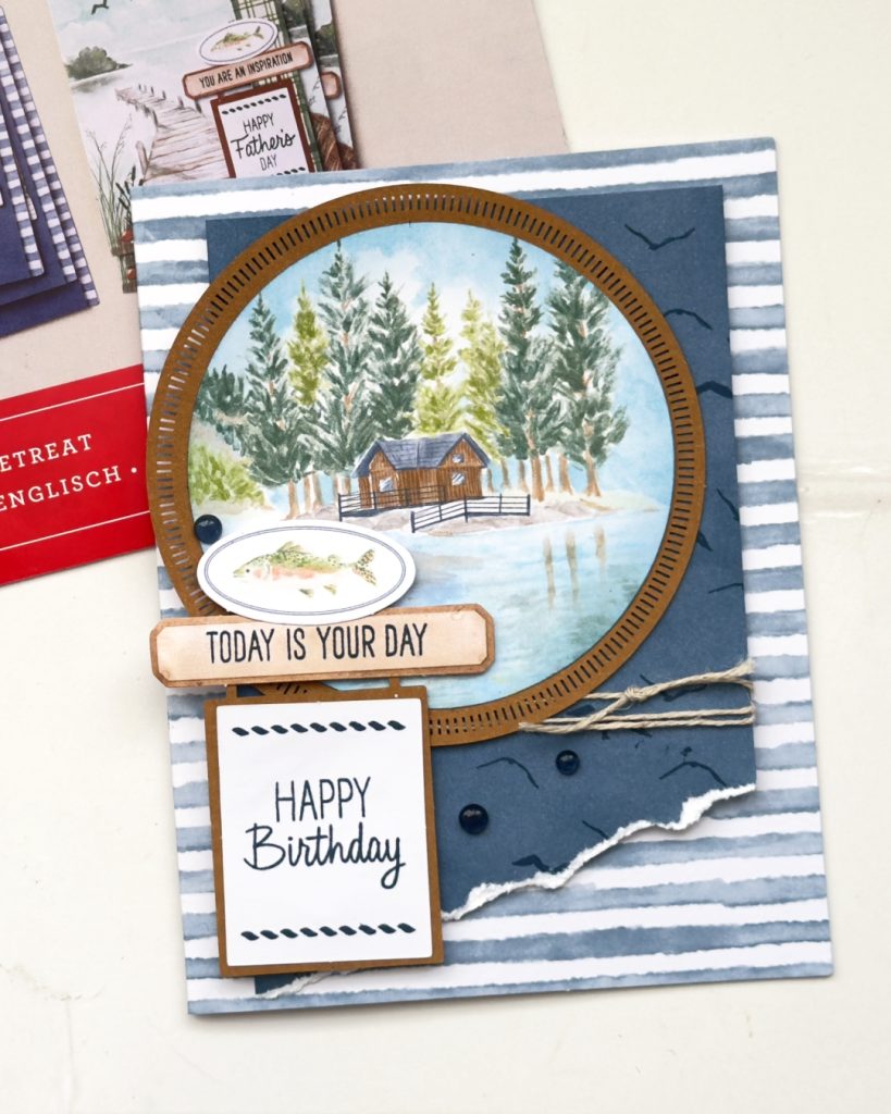 Handmade masculine birthday card featuring a circular lakeside cabin scene with pine trees, layered over striped patterned paper with twine, fishing embellishments, and “Happy Birthday” sentiment in blue and earthy tones.